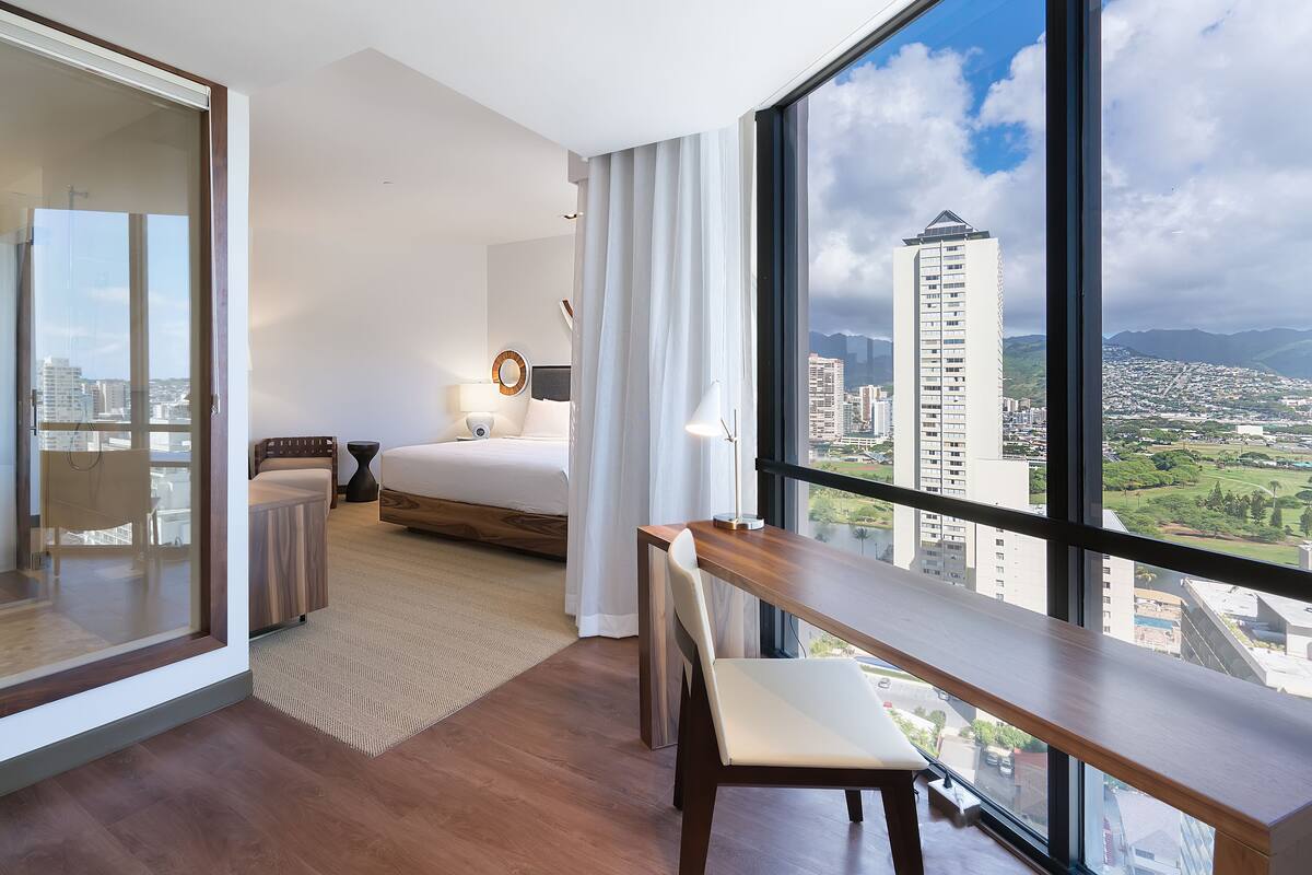 A modern hotel room with a large window, a bed, a desk, and a view of the cityscape.