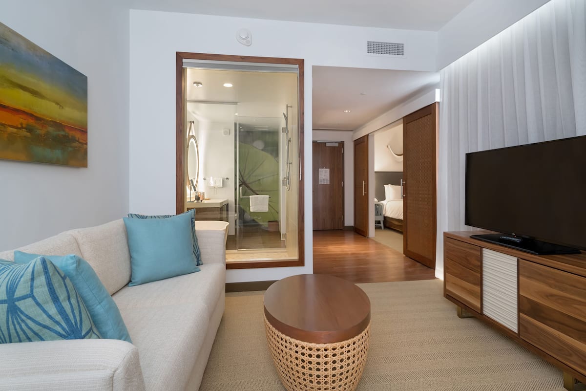 A modern living room with a sofa, a round coffee table, a television on a wooden cabinet, and a glass door leading to a bathroom.
