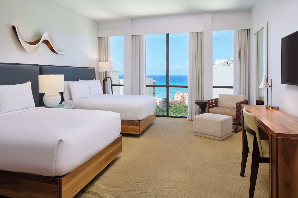 A hotel room with two beds, a desk, a chair, and a large window offering a view of the city.