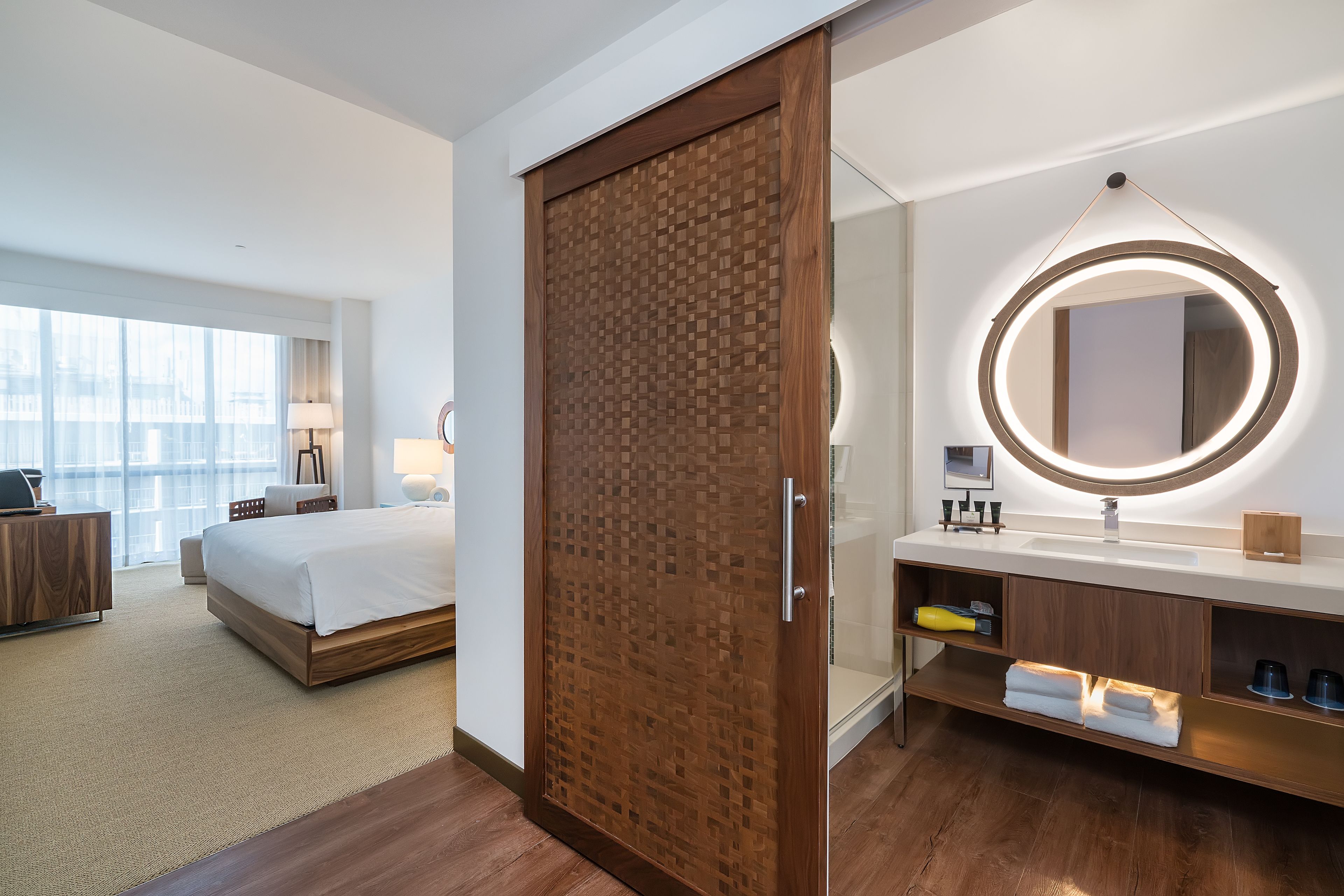 Ein modernes Hotelzimmer mit einem großen Bett, einem Holzschrank, einem runden Spiegel mit Beleuchtung, einem Badezimmer mit Waschbecken und Regalen sowie Blick auf die Stadt durch große Fenster.