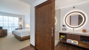 Una habitación de hotel moderna con una cama grande, un armario de madera, un espejo redondo con luz, un baño con lavabo y estanterías, y vistas a la ciudad a través de grandes ventanales.