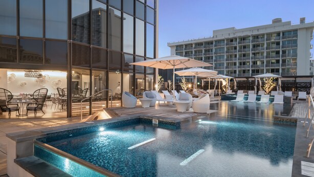 A rooftop infinity pool with lounge chairs and umbrellas, surrounded by modern buildings.