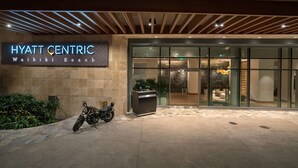 The exterior of the Hyatt Centric Waikiki Beach hotel with a motorcycle parked in front, a reception desk, and a modern entrance.