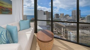 Habitación de hotel moderna con un ventanal grande que ofrece vista a la ciudad, un sofá cómodo con cojines azules y una mesa redonda de madera.