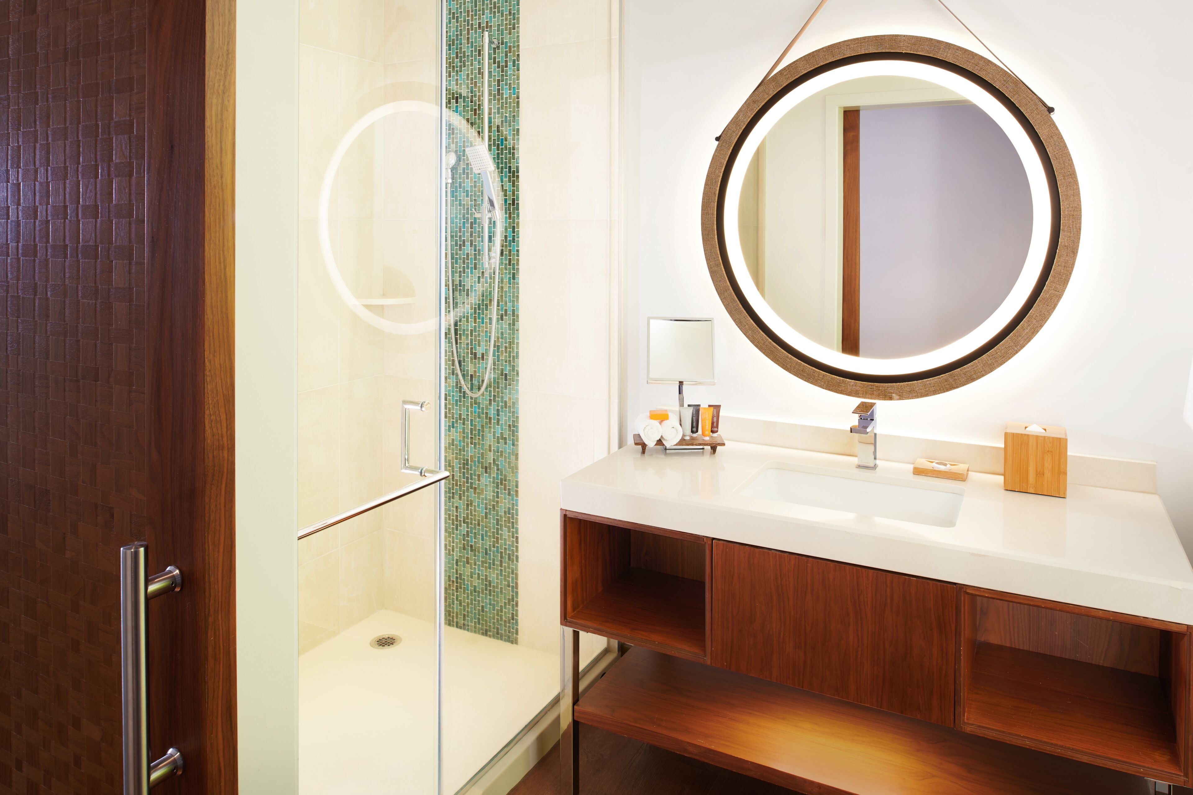 A modern bathroom with a glass shower enclosure, a round mirror, and a wooden vanity.