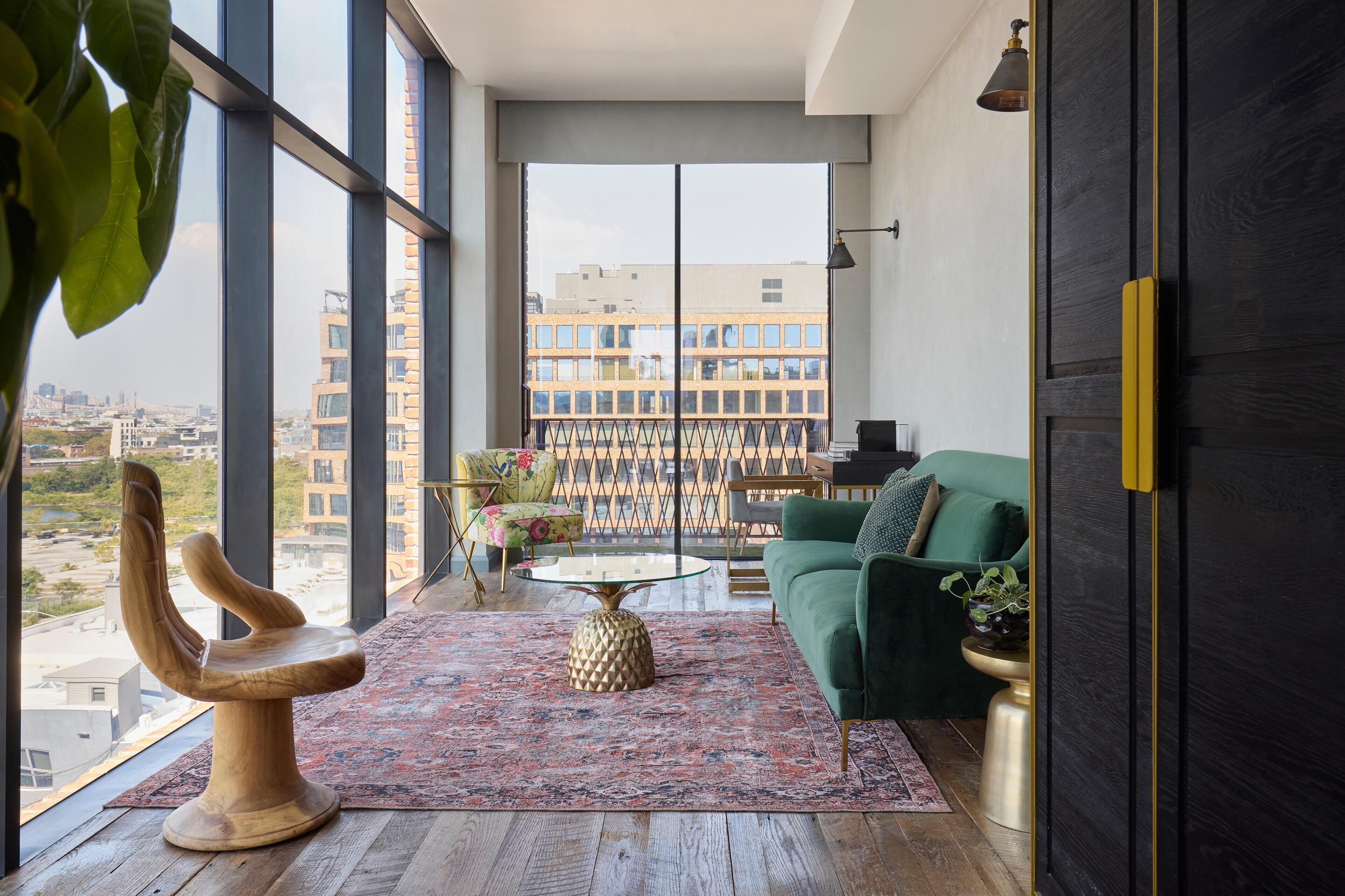 A modern living room with a large window, a green sofa, a wooden chair, and a patterned rug.