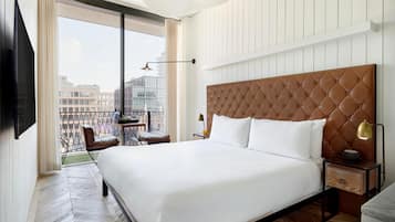 A hotel room with a large bed, a TV mounted on the wall, a balcony with a view of the city, and a modern light fixture.