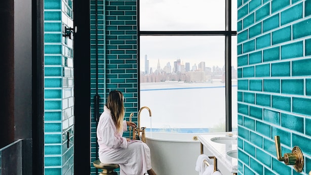 A bathroom with a large bathtub, a window with a city view, and teal brick walls.