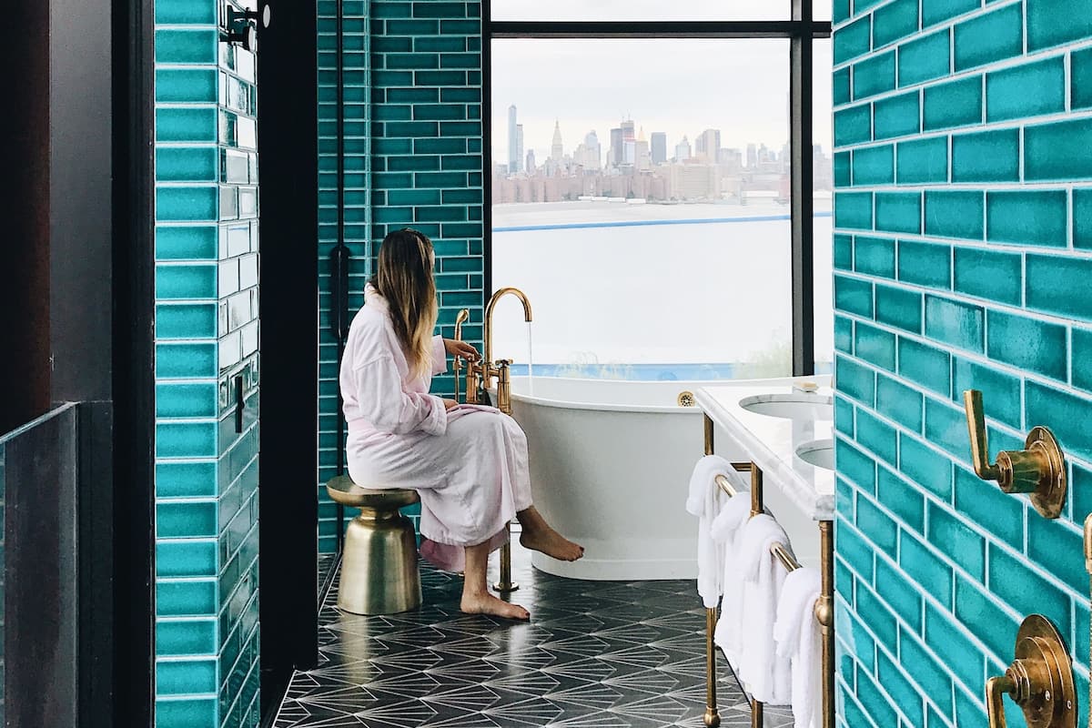 Un baño con una bañera grande, una ventana con vistas a la ciudad y paredes de ladrillo color aguamarina.