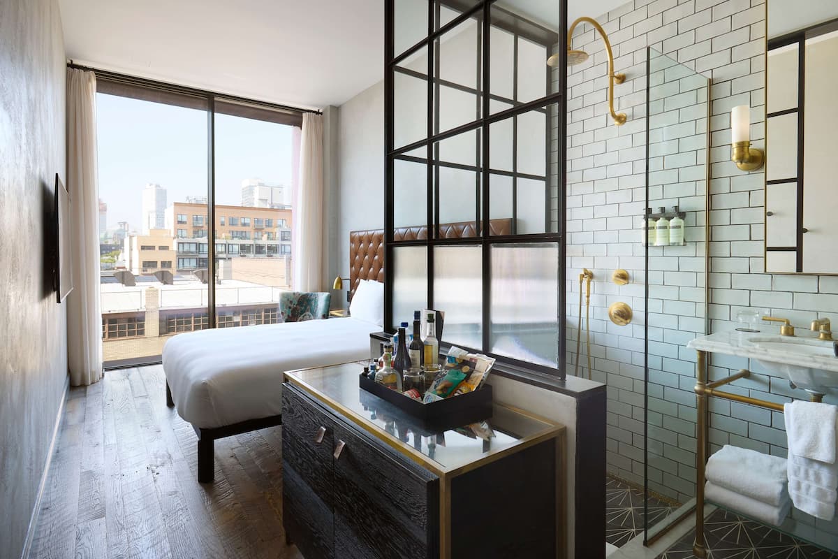 Une chambre d’hôtel moderne dotée d’un grand lit, d’une commode à plateau en verre, d’une salle de bain avec une douche à parois de verre et offrant une vue sur la ville.