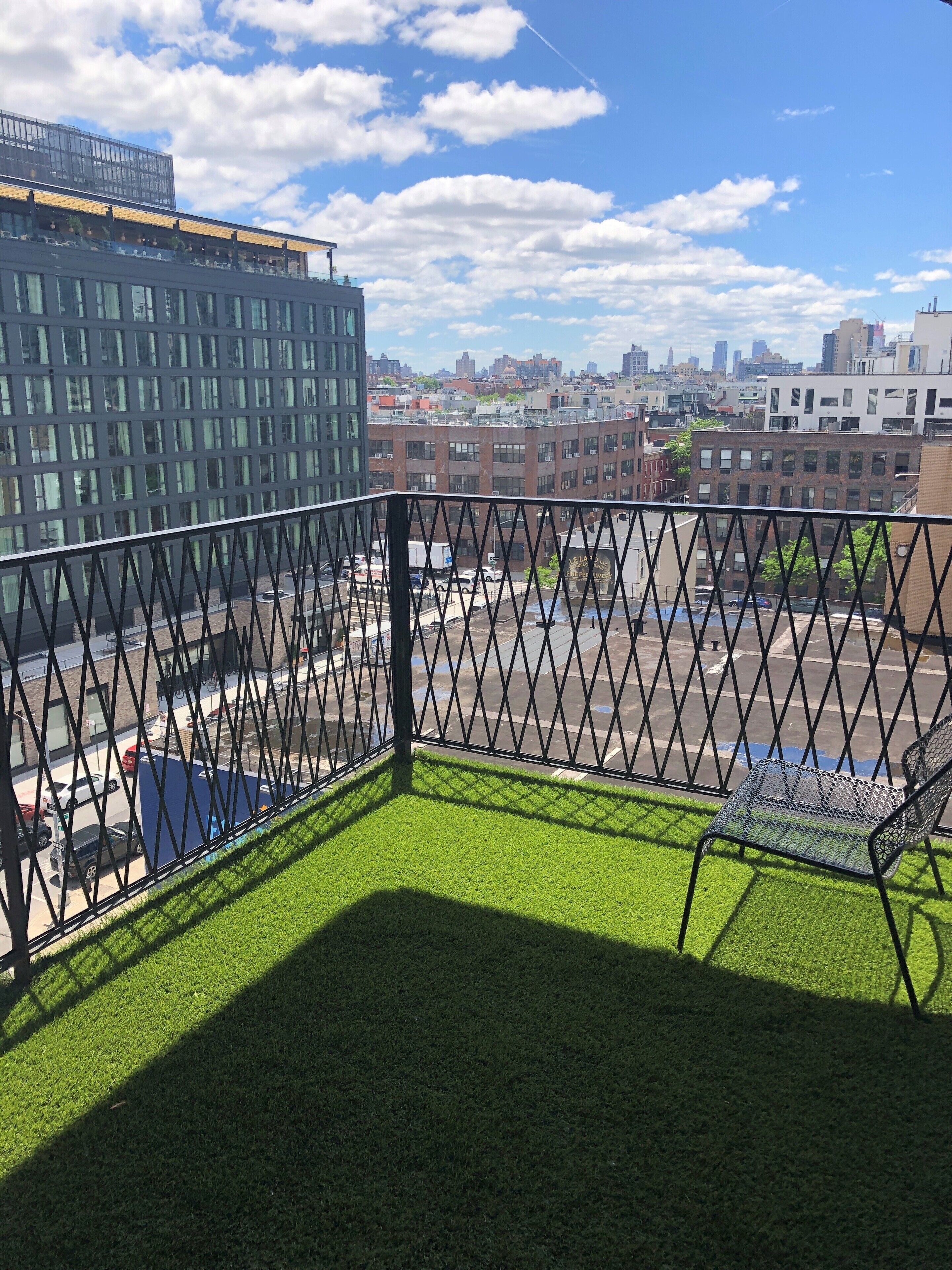 A balcony with artificial grass, a metal railing, and a view of a cityscape with buildings and a parking lot.