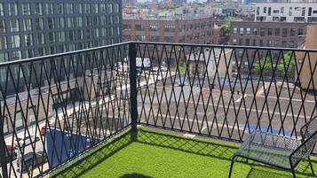 A balcony with artificial grass, a metal railing, and a view of a cityscape with buildings and a parking lot.