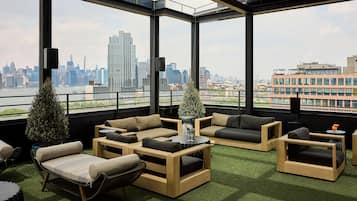A rooftop lounge with a city view, furnished with sofas and tables, and a small Christmas tree.
