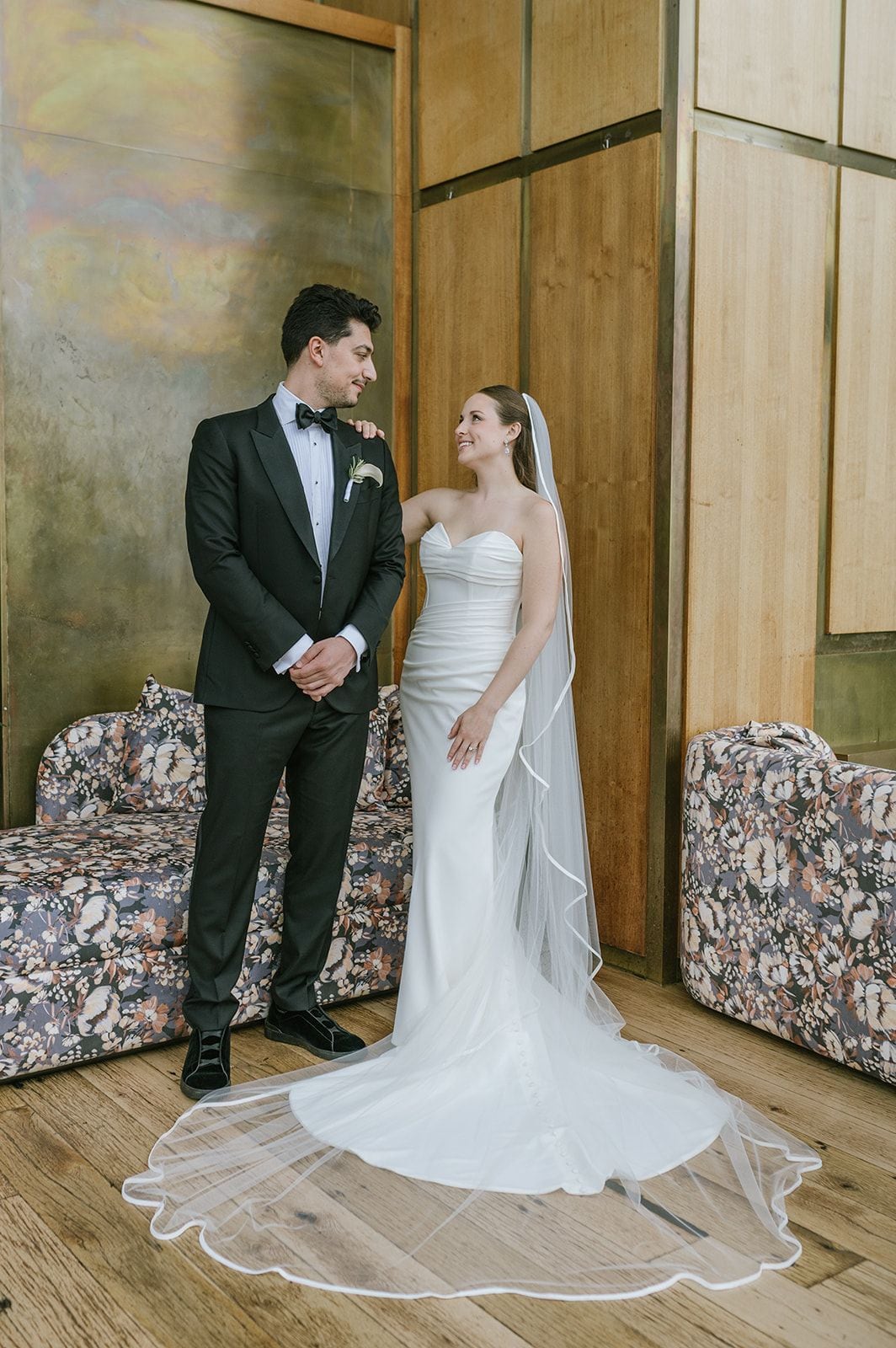 Una pareja vestida de novios parada en una habitación con paneles de madera y sofás de estampado floral.