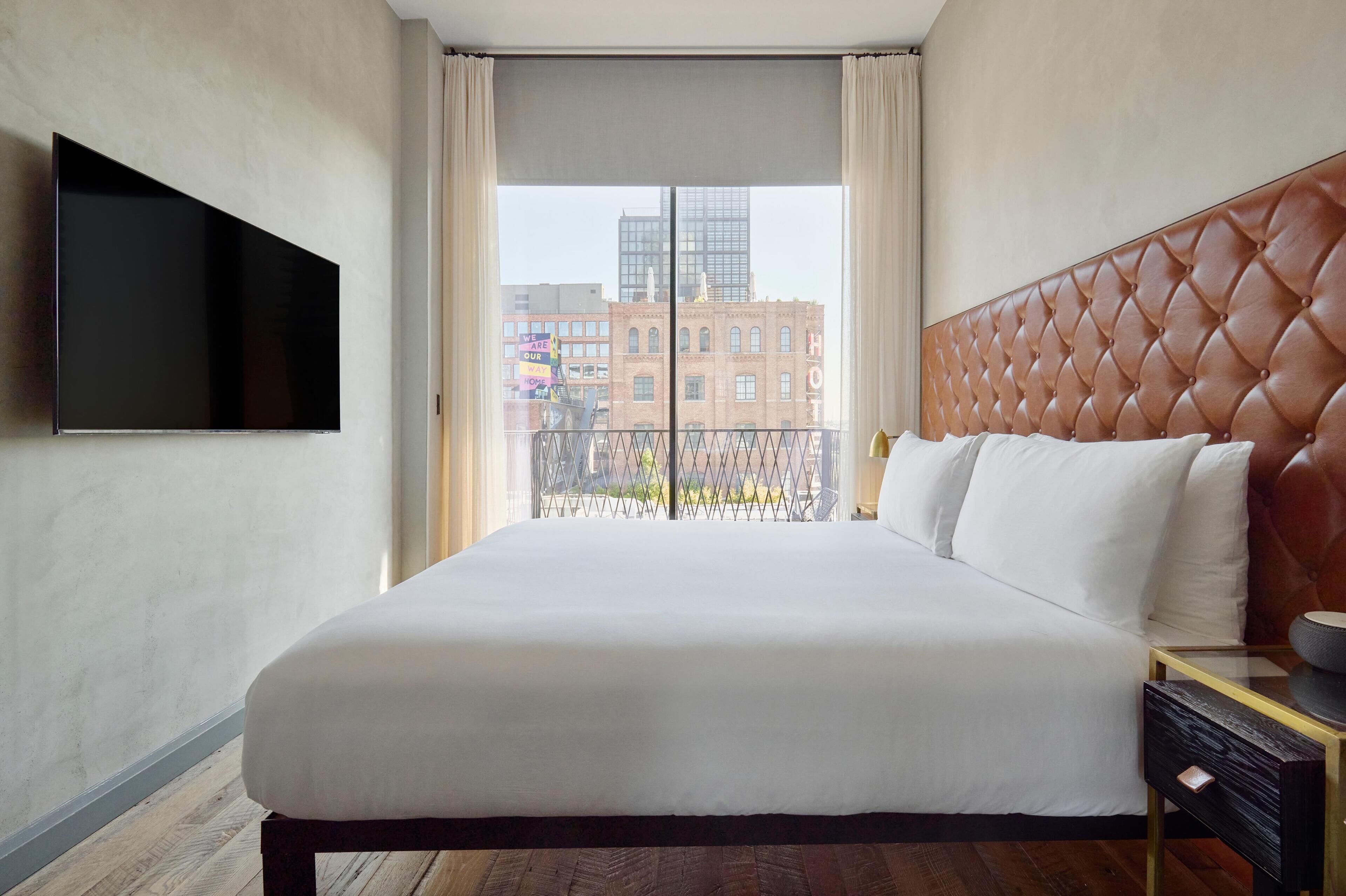 A hotel room with a large bed, a flat-screen TV mounted on the wall, and a view of the city through a window.