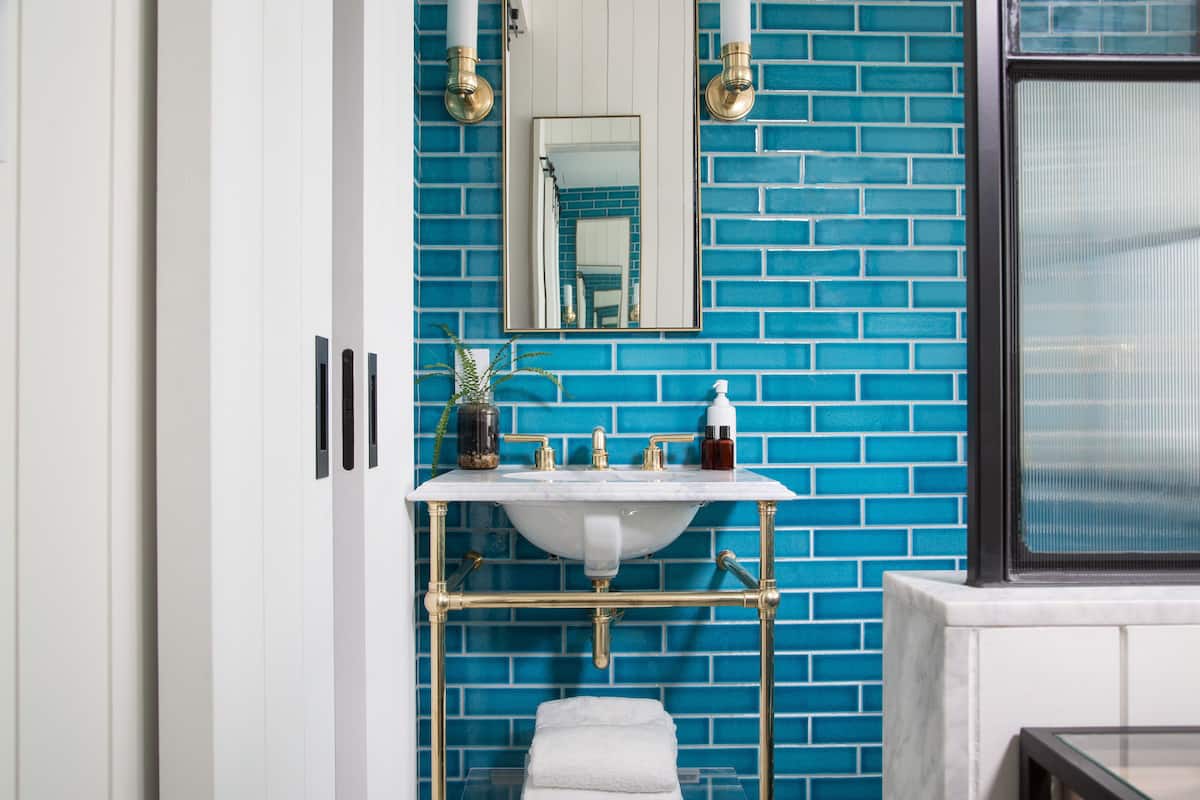 Un baño con lavamanos blanco, paredes con azulejos azules, un espejo y una ventana.