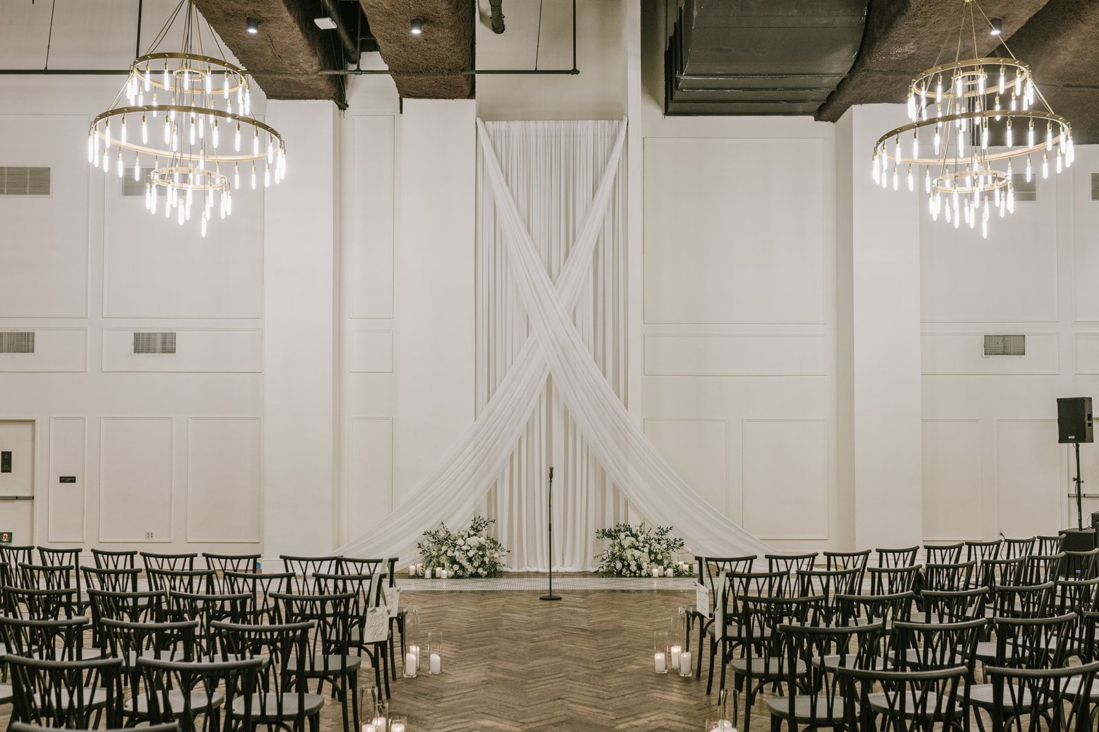 A venue with a central aisle flanked by rows of chairs, a stage with draped fabric, and chandeliers.
