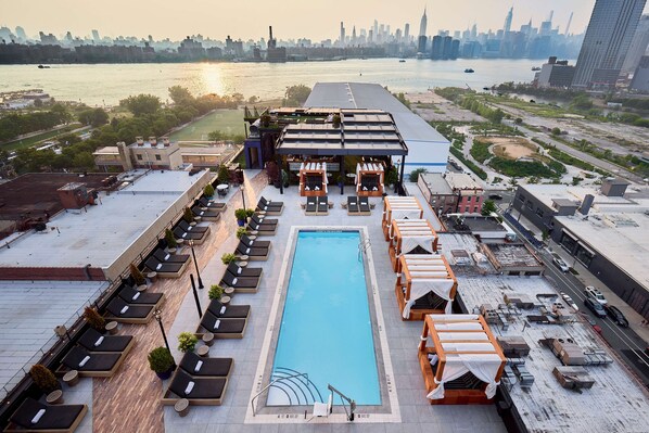 Una piscina en la azotea con tumbonas y una zona de estar cubierta, con vistas a un horizonte urbano y un río.