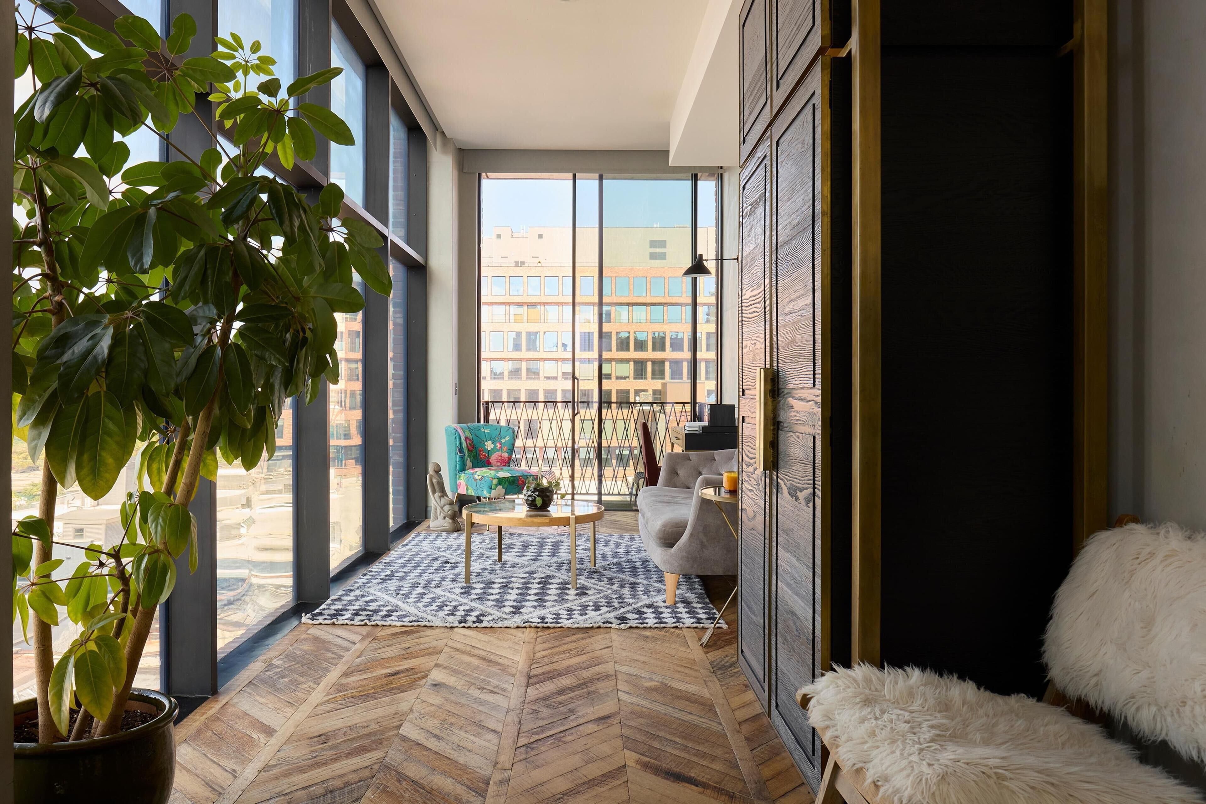 A modern interior with large windows, a herringbone floor, a potted plant, a chair, a sofa, and a fluffy white pouf.