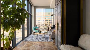 A modern interior with large windows, a herringbone floor, a potted plant, a chair, a sofa, and a fluffy white pouf.