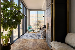 A modern interior with large windows, a herringbone floor, a potted plant, a chair, a sofa, and a fluffy white pouf. - Arlo Williamsburg (Brooklyn)