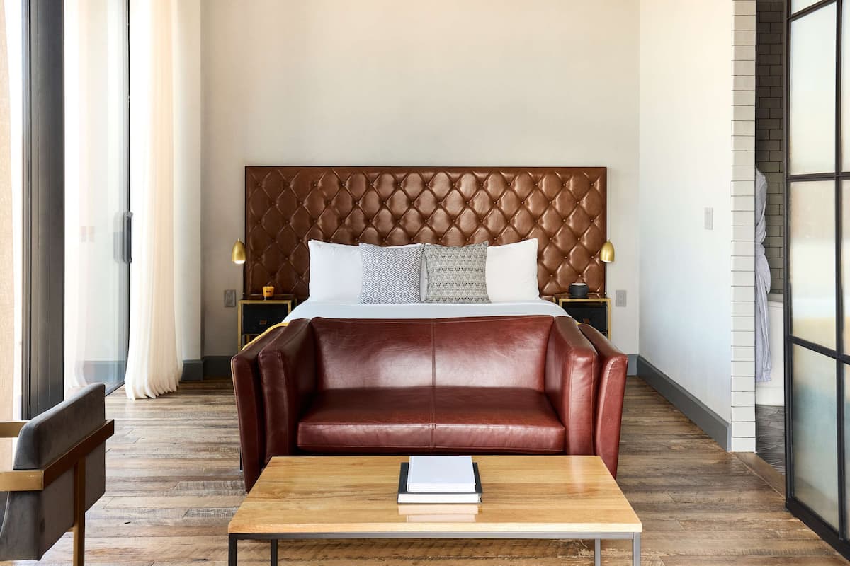 Une chambre d’hôtel avec un grand lit, un canapé en cuir, une table basse en bois et une vue sur l’extérieur, visible à travers une porte vitrée.
