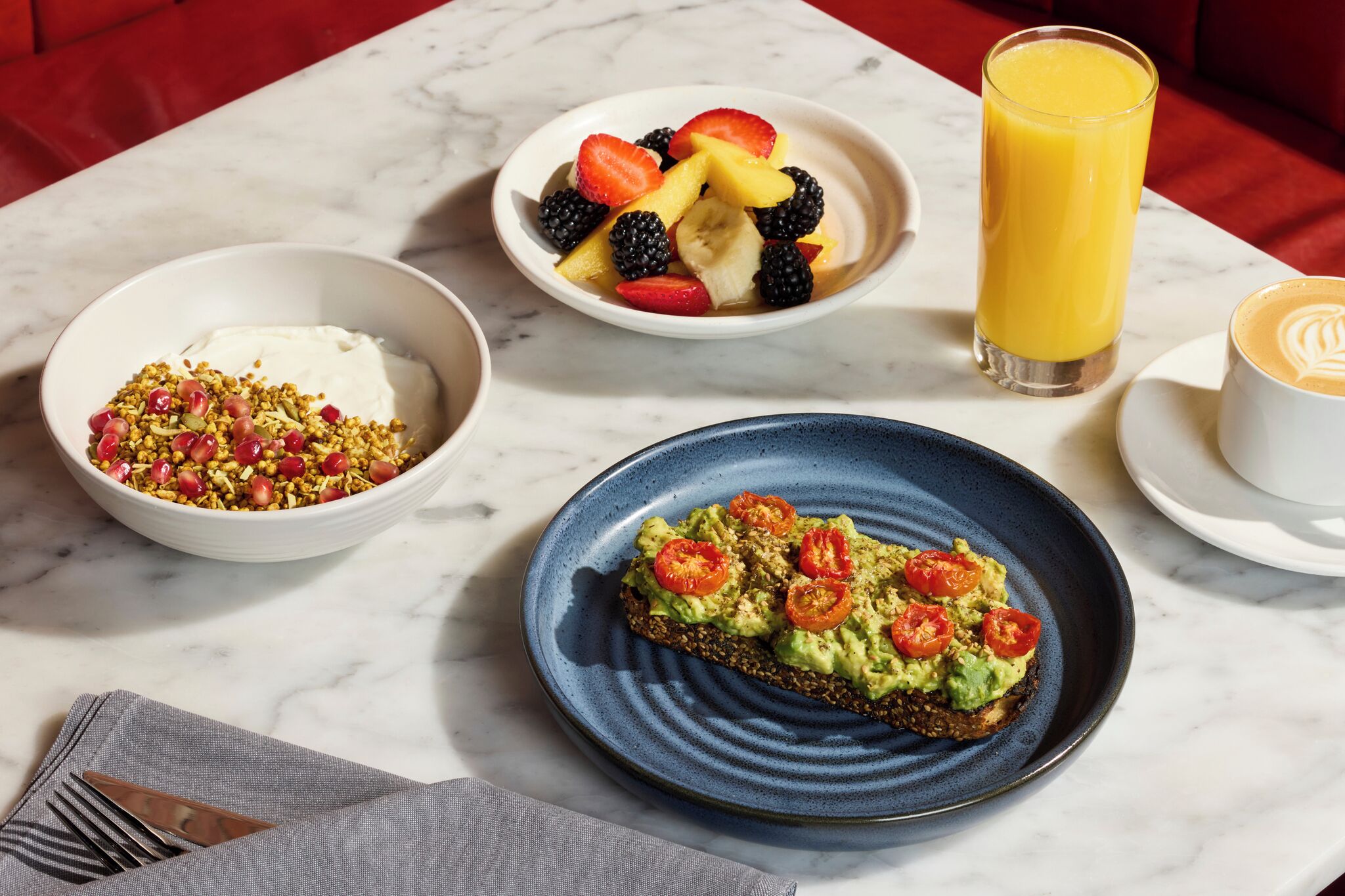 Een ontbijttafel met een bakje yoghurt met granola, een bakje gemengd fruit, een glas jus d'orange en een bord met avocado toast.