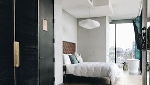 A modern bedroom with a large bed, a wooden herringbone floor, a black wardrobe, and a city view through a large window.