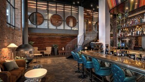 A bar area with a herringbone floor, a bar counter, and blue bar stools. There are round mirrors on the wall, a staircase, and a brick wall.