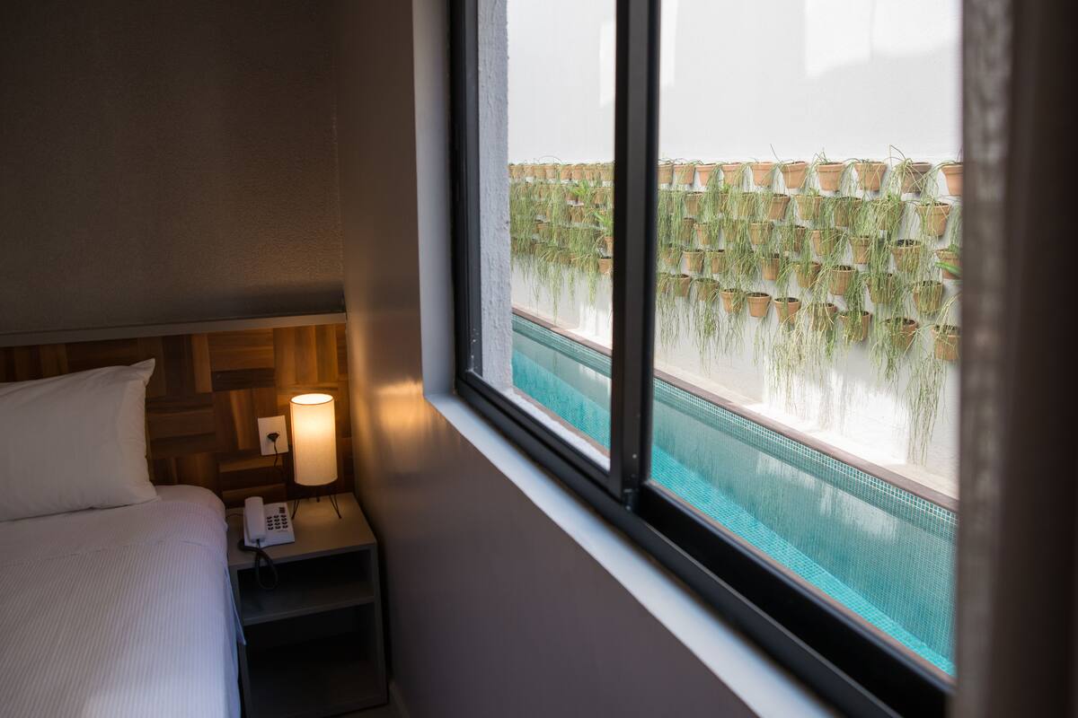 Habitación de hotel con cama, lámpara de noche y vista a una piscina con plantas en macetas.