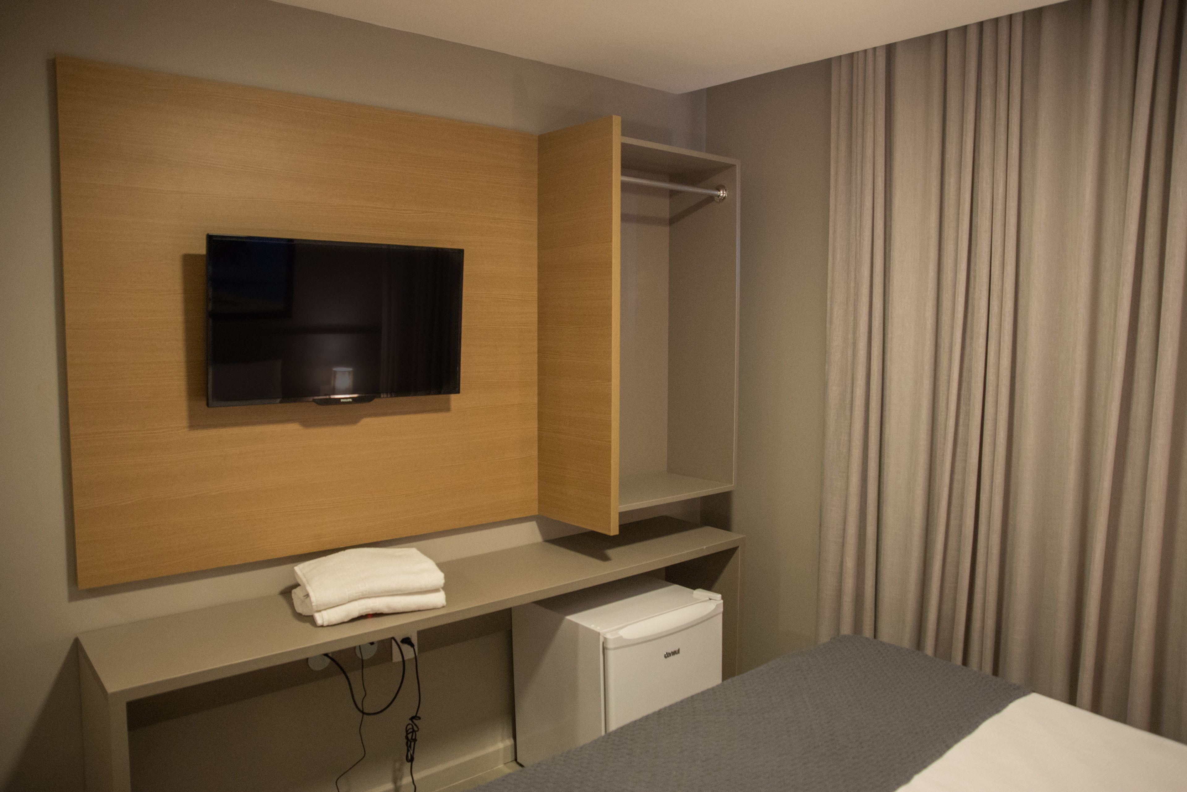 A hotel room with a flat-screen TV mounted on a wooden panel, a small white minibar, and a grey bed.