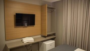 A hotel room with a flat-screen TV mounted on a wooden panel, a small white minibar, and a grey bed.