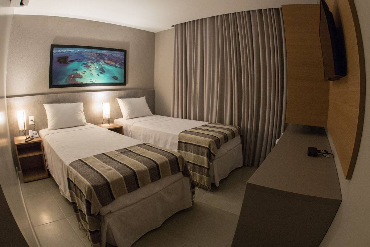 Habitación de hotel con dos camas, un televisor montado en la pared y un ventanal con cortinas.
