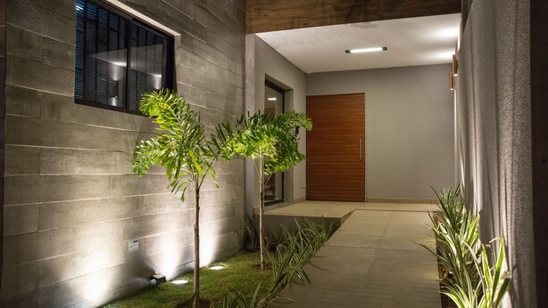 A modern building exterior with a concrete facade, a wooden door, and a well-lit pathway with small trees and plants. - Amenit Hotel (Maceió)