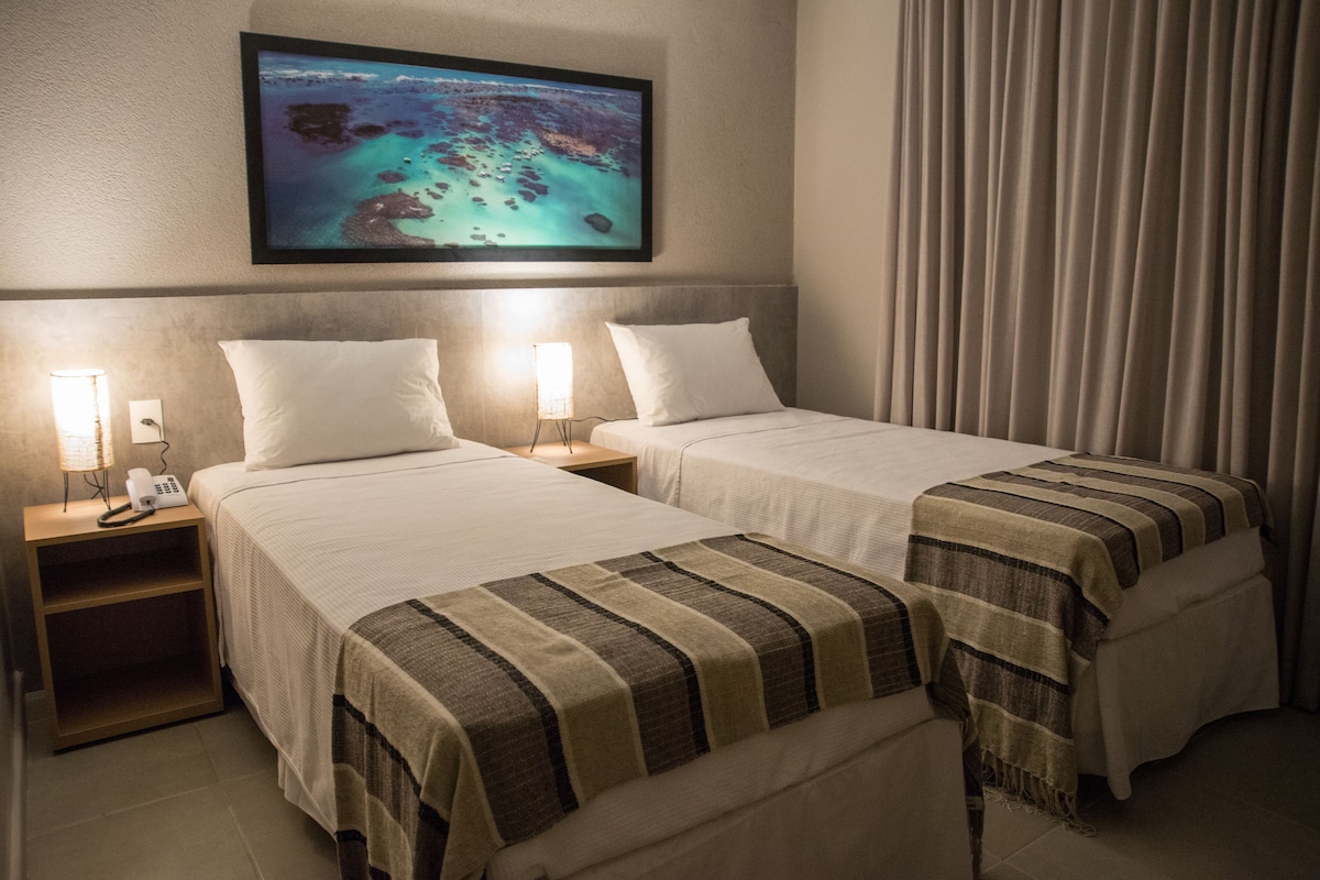 A hotel room with two beds, a nightstand with a phone, a lamp, and a painting of a beach scene.