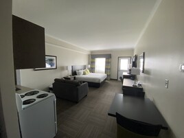 Habitación de hotel con cama, escritorio, silla, una pequeña cocina americana con estufa y ventana con cortinas.