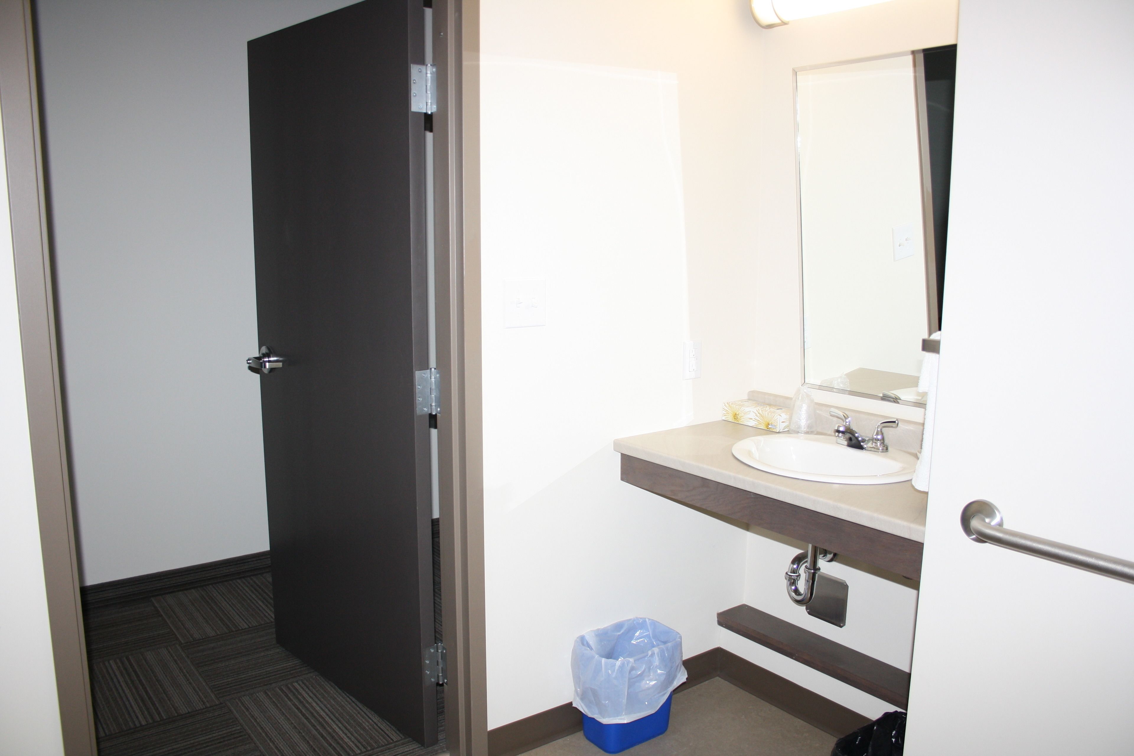 Un baño de hotel con lavamanos blanco, un espejo, un cubo de basura y una puerta.