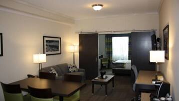 A hotel room with a living area, a dining table, a sofa, and a bed. There are lamps on either side of the sofa and a picture on the wall.