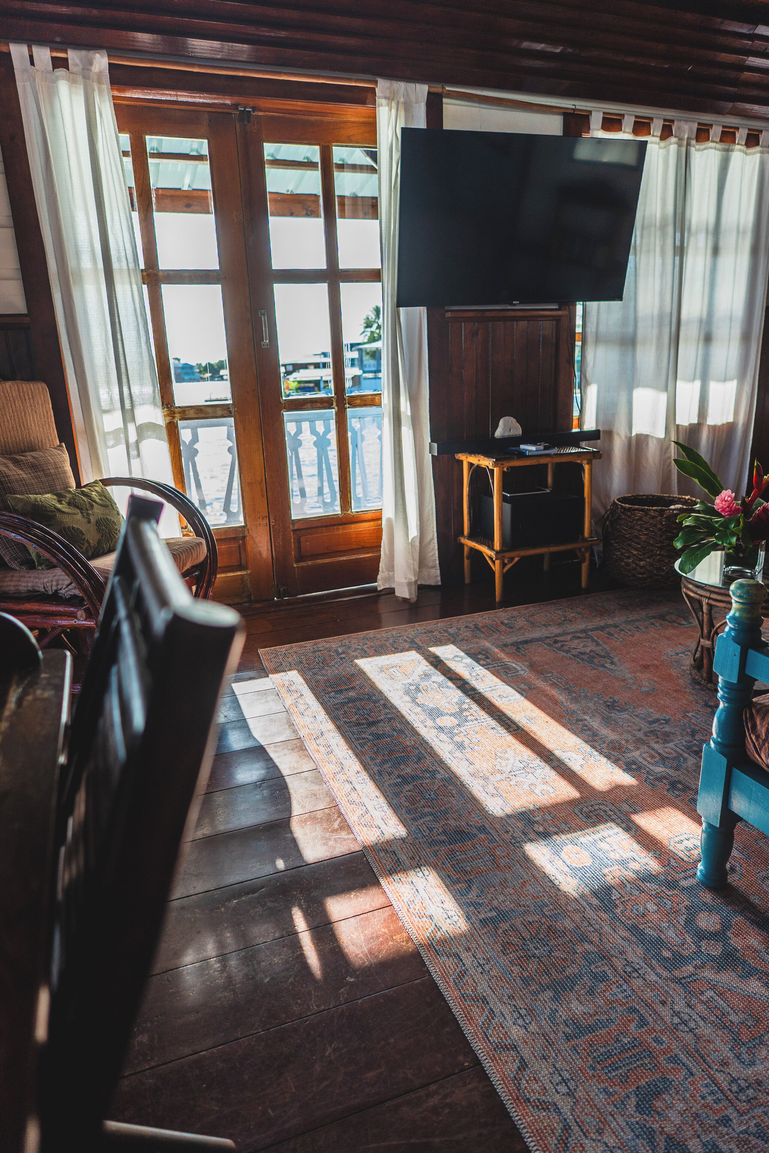 Una sala de estar con piso de madera, un televisor de pantalla plana grande montado en la pared, una silla de mimbre, una mesa de centro de madera y vistas a través de una puerta de cristal.