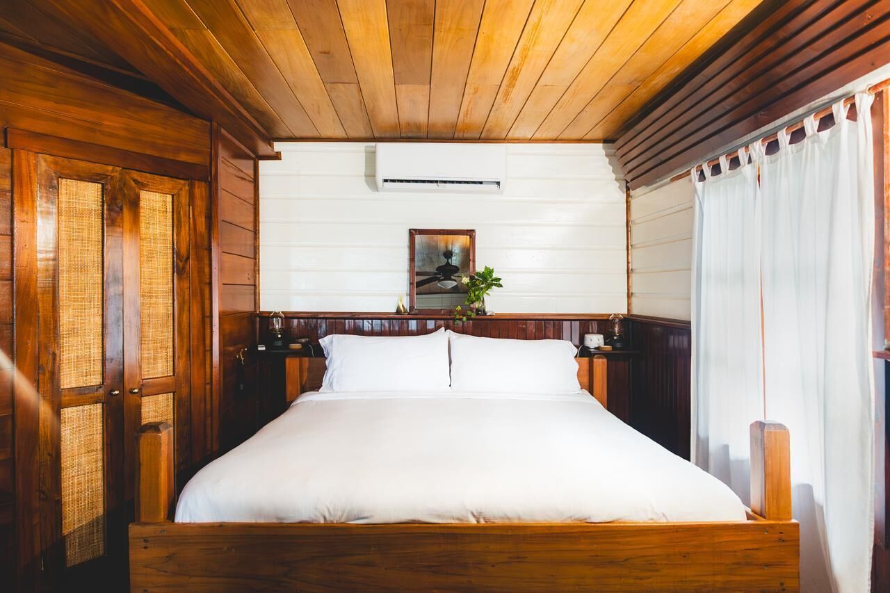 Una cama con estructura de madera y ropa de cama blanca, un aire acondicionado montado en la pared y un cuadro enmarcado encima.