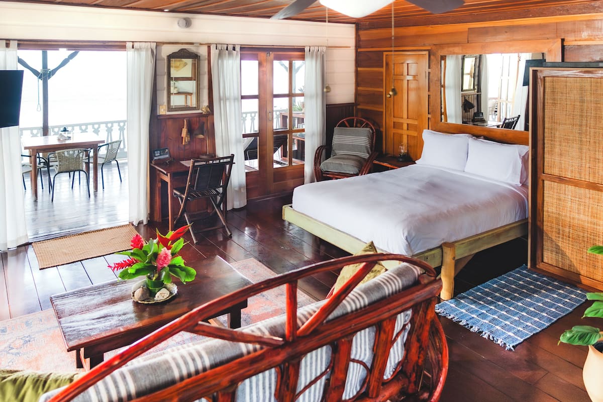 A wooden-floored bedroom with a bed, a chair, a table, and a balcony with a table and chairs.