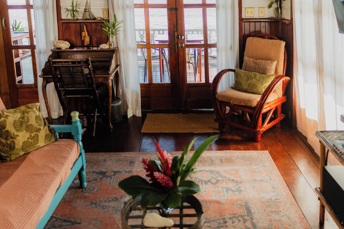A cozy room with wooden flooring, a sofa, a rocking chair, a table with a plant, and a view through a large window.