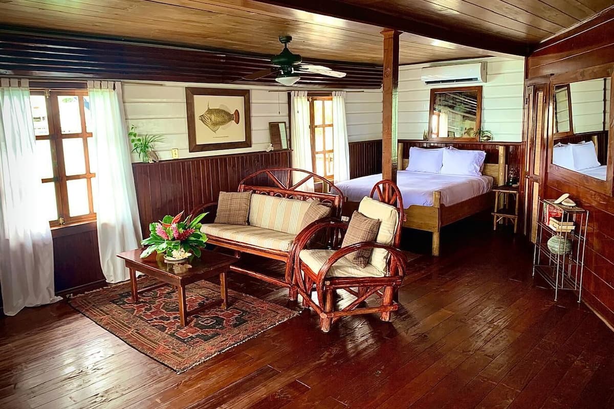 A wooden-floored room with a bed, two wicker chairs, a small table with a plant, and a ceiling fan.