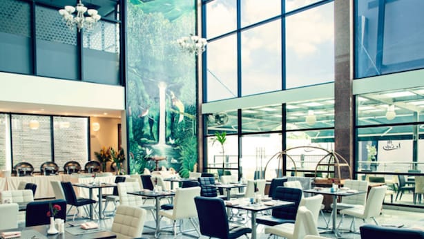 A spacious indoor area with tables and chairs arranged for dining, large windows allowing natural light, and a mural depicting a natural landscape on the wall. - Laguna Hotel (Port Moresby)