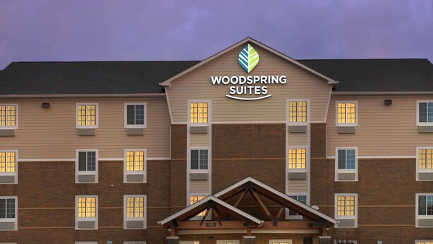 Un hotel con un cartel llamativo de "Woodspring Suites", varias ventanas y un estacionamiento con un automóvil.