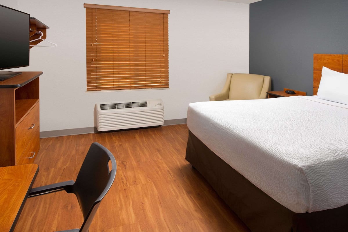 A hotel room with a bed, a desk with a chair, a television, a window with blinds, and an air conditioning unit.