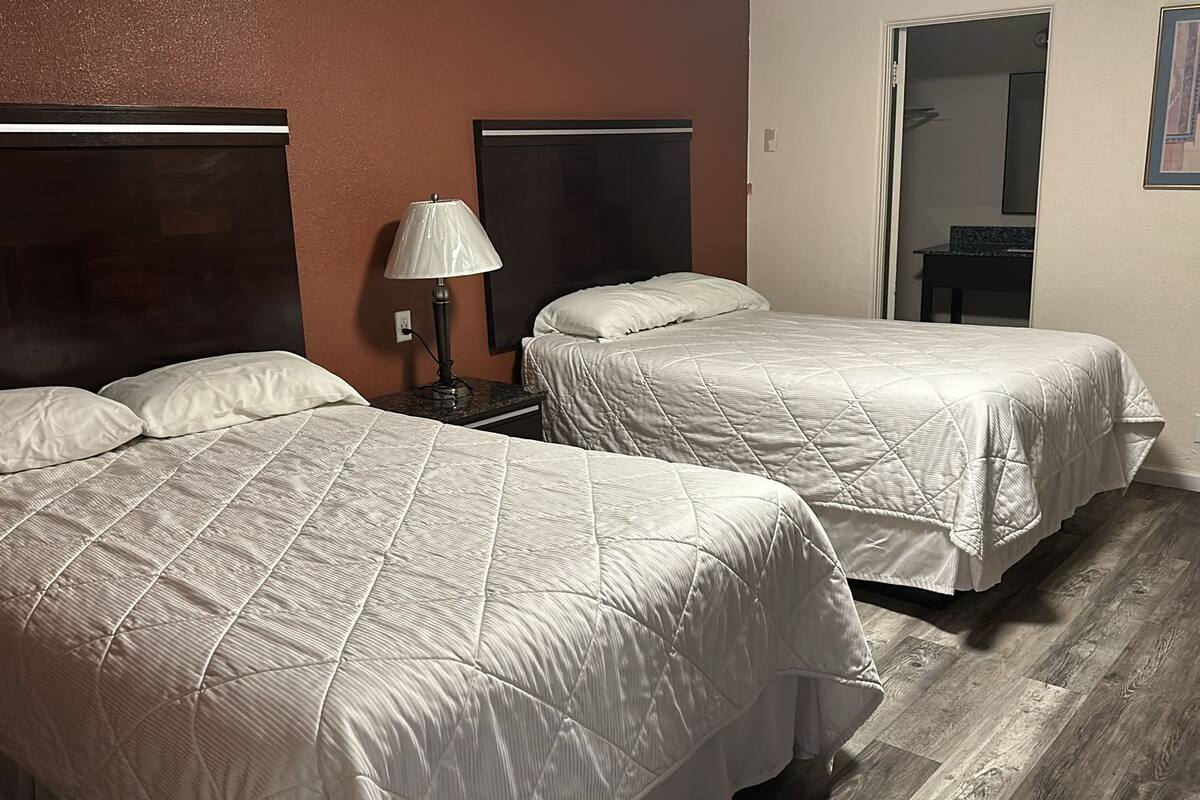 A hotel room with two beds, a nightstand, a lamp, and a door leading to a bathroom.