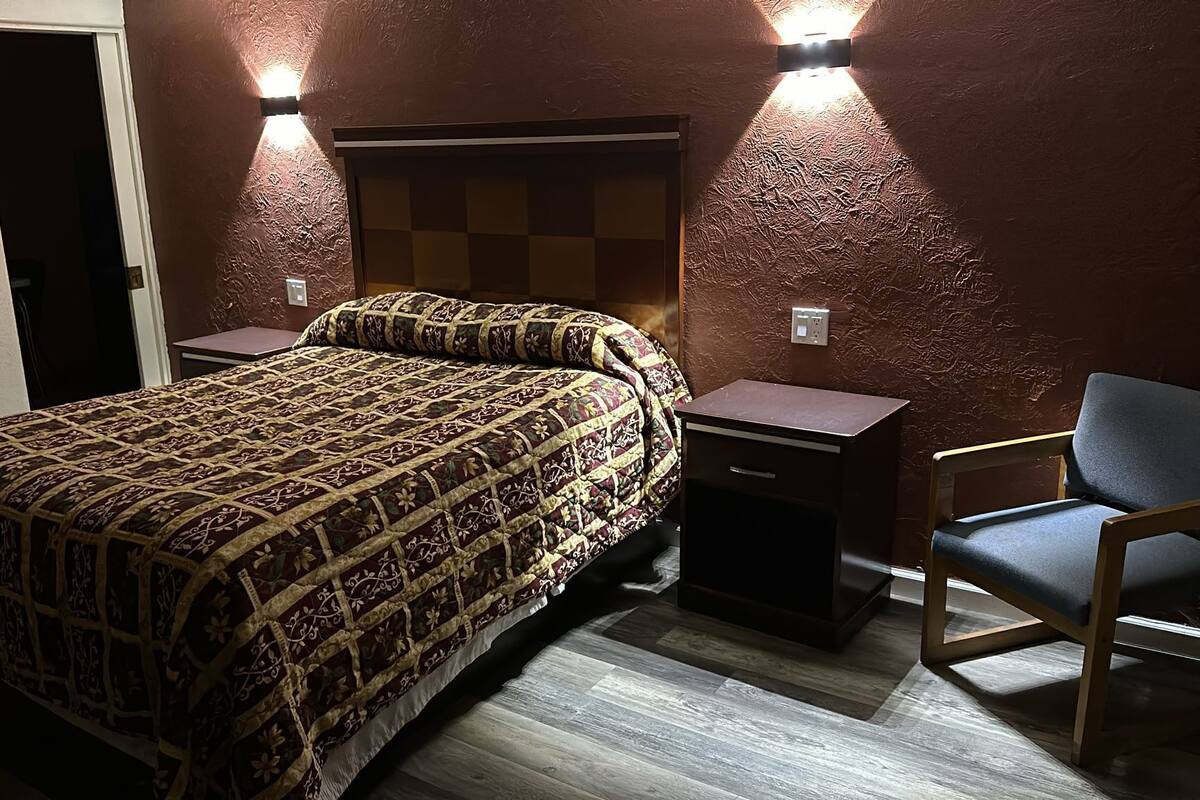 A bedroom with a patterned bedspread, a nightstand, a chair, and two wall-mounted lights.