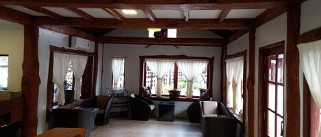 A room with wooden beams, a tiled floor, wicker furniture, and large windows with curtains.