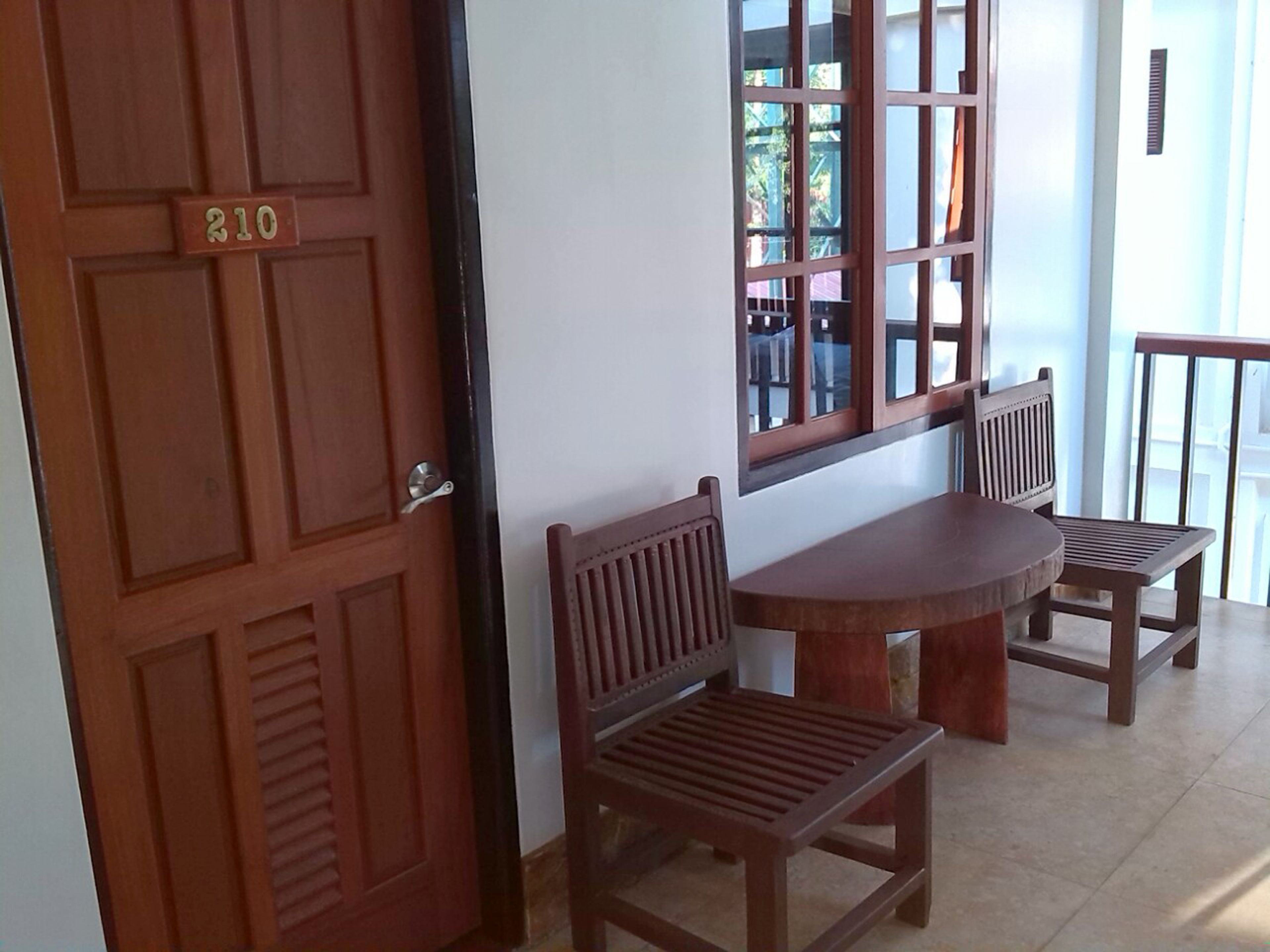 Una puerta de madera con una placa numérica, una ventana que ofrece vistas a muebles de exterior, y una mesa redonda con dos sillas de madera.